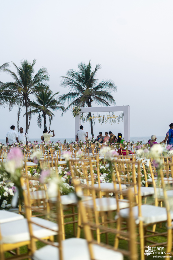 Wedding ceremony seating area on the beach with gold chairs, flowers, and palm trees in background.