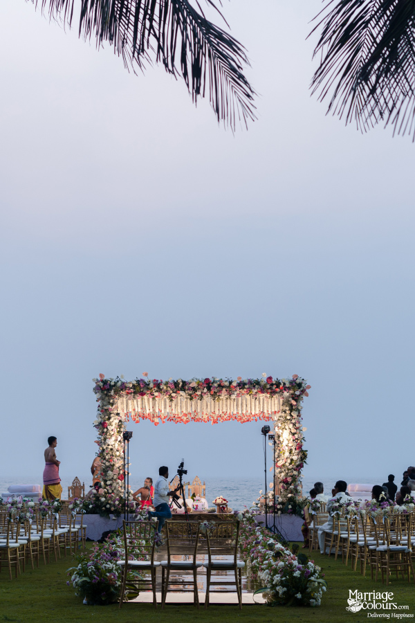Floral beach wedding mandap with gold chairs, overlooking the ocean under a pale evening sky.