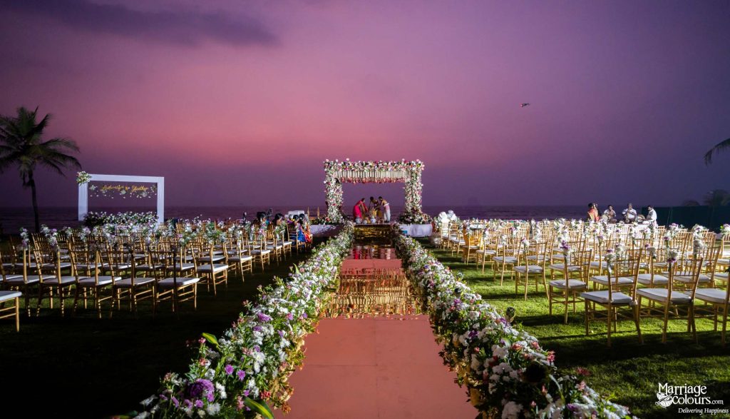 Romantic beach wedding ceremony with a floral aisle, gold chairs, and mandap against a purple sky.
