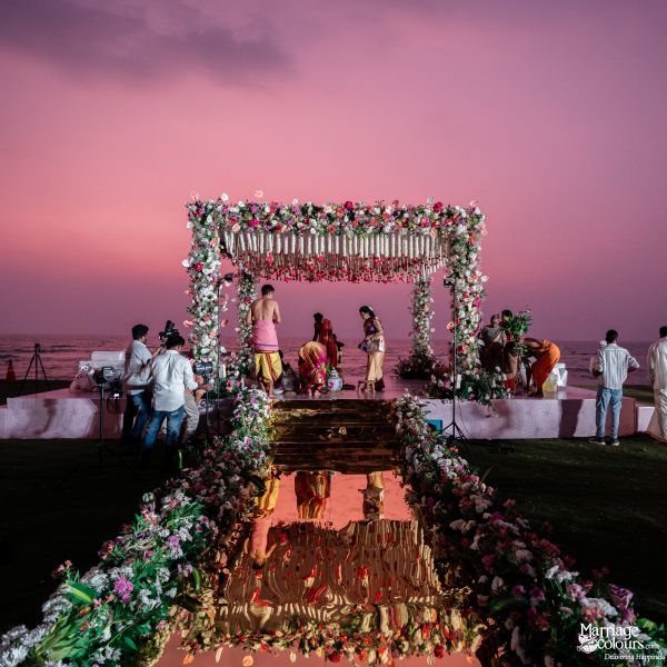 Indian wedding ceremony at sunset with a floral mandap, mirrored aisle, and a dramatic pink sky.