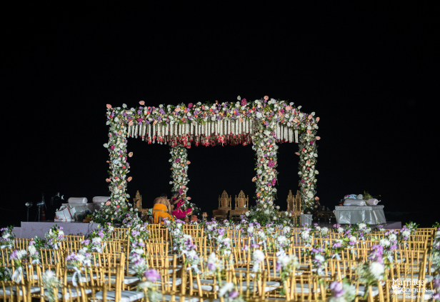 Night-time Indian wedding ceremony with a richly decorated floral mandap and rows of gold chairs.