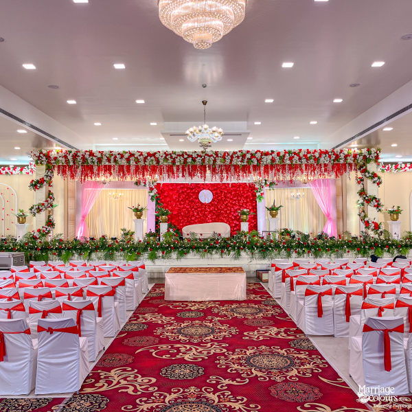 Wedding reception hall with red flower stage, chandelier, and white chairs with red sashes.