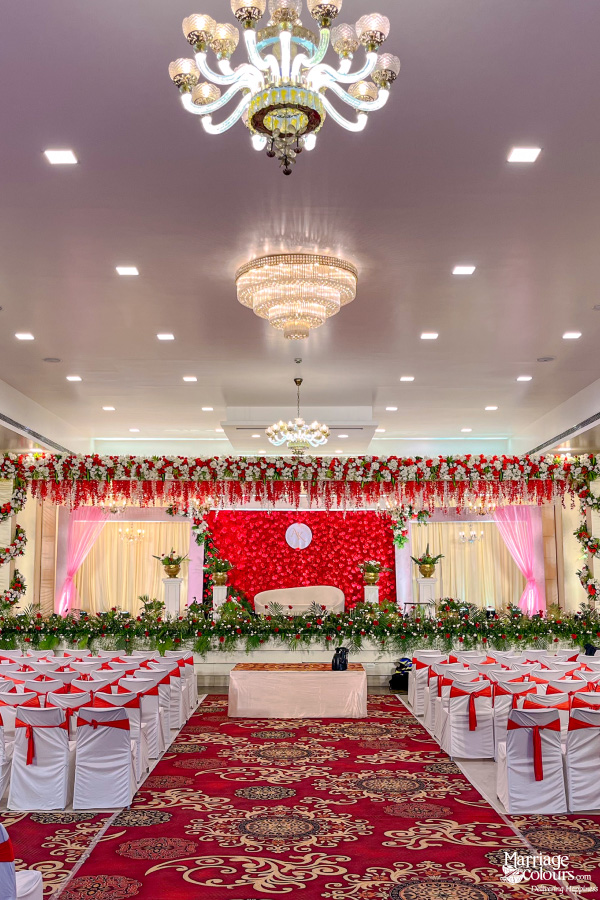 Wedding reception hall with red flower stage, tiered chandeliers, and white chairs with red sashes.