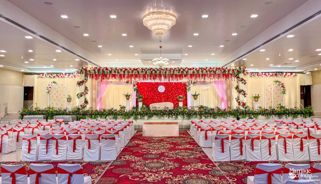 Wedding reception hall with red flower stage, chandelier, and white chairs with red sashes.