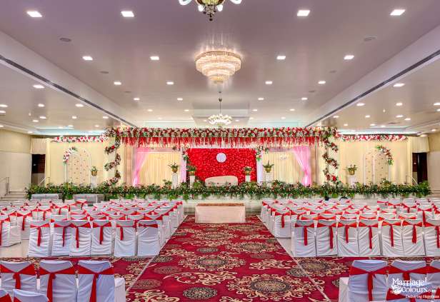 Wedding reception hall with red flower stage, tiered chandeliers, and white chairs with red sashes.