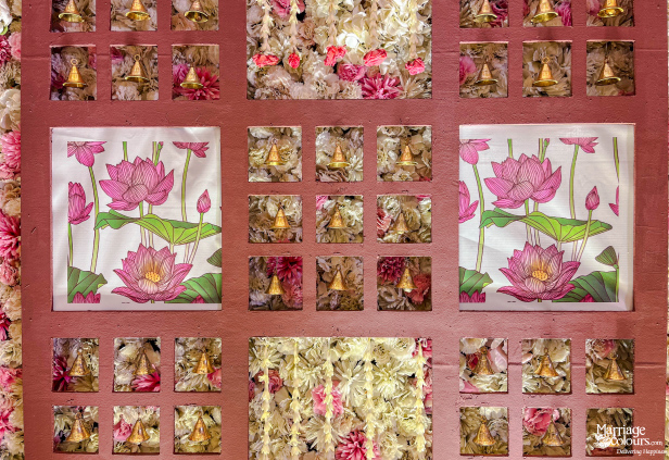 Wedding decor wall panel with pink lotus art, small golden bells, and clusters of fresh flowers.