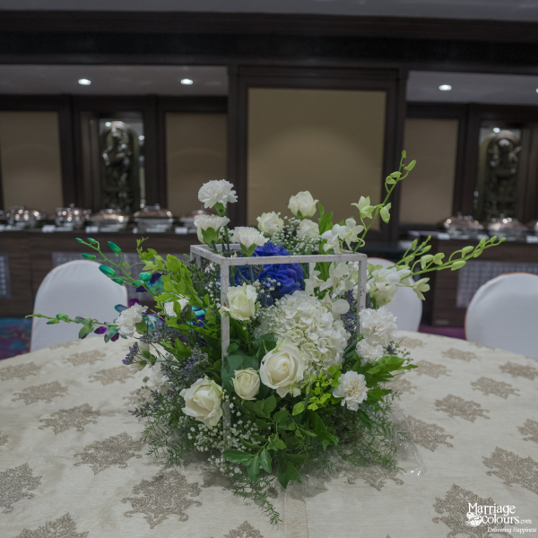 Modern table centerpiece featuring white roses, blue blooms, and green foliage in a geometric wire frame.