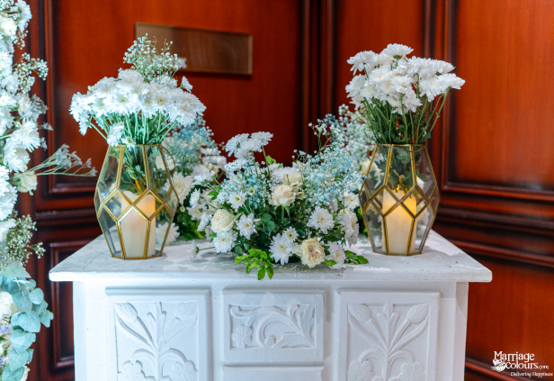 White mums and baby's breath arranged on a carved white table with geometric glass lanterns.