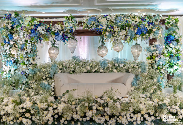 Elaborate wedding stage with a white sofa, surrounded by blue and white floral garlands and crystal lights.