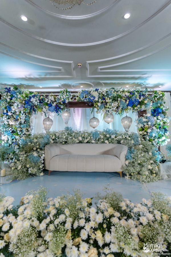 Wedding stage with floral backdrop, chandelier, lantern lights, and white sofa. 📝 Description A dreamy wedding stage featuring white-blue flowers, glowing lanterns, chandelier lighting, and a plush sofa set against a lush floral wall. Keep them coming—I’m loving the elegance in these setups!