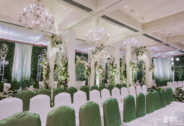 Wedding ceremony seating arrangement with white and green chairs beneath ornate floral arches and chandeliers.