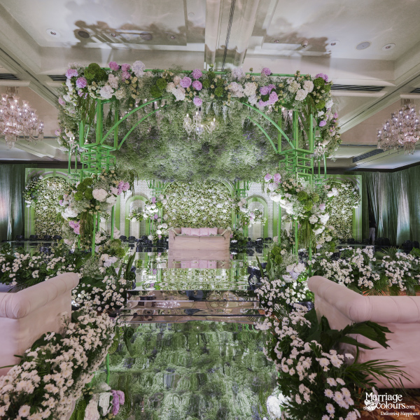 Highly decorative, luxurious wedding stage with extensive white florals, vibrant greenery, and a mirrored aisle.