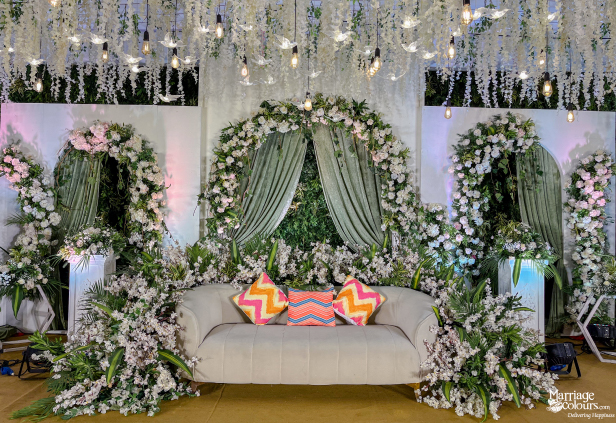 Elegant indoor stage with a white sofa, floral arches, sage green drapes, and hanging wisteria.