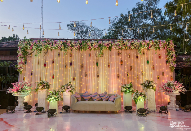 Wedding stage featuring pink drapery, floral borders, small lanterns, and a neutral loveseat for the couple.