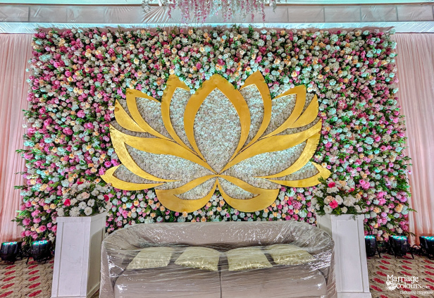 Wedding stage backdrop with a large golden lotus, dense flower wall, and a plastic-covered sofa.