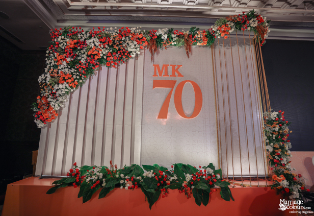 Close-up of a stage backdrop featuring an illuminated orange 'MK 70' sign with floral trim.