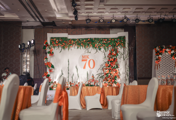 White event backdrop with orange 'MK 70' logo, bright florals, and vine decorations.