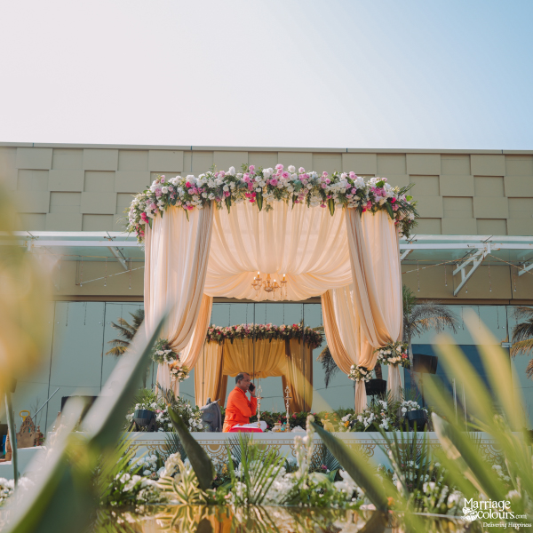 Outdoor wedding mandap with peach fabric drapes, a floral roof, and a priest performing rituals.