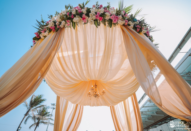 Close-up of a wedding mandap canopy with peach drapes, vibrant flowers, and blue sky.