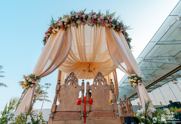 Outdoor wedding mandap with peach fabric drapes, a floral roof, and ornate wooden chairs.