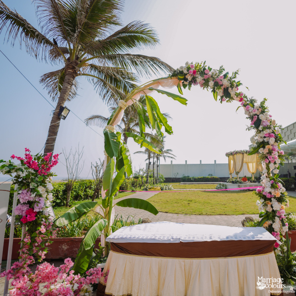 Outdoor ceremony table with a banana leaf and floral arch on a grassy, palm tree-lined lawn.