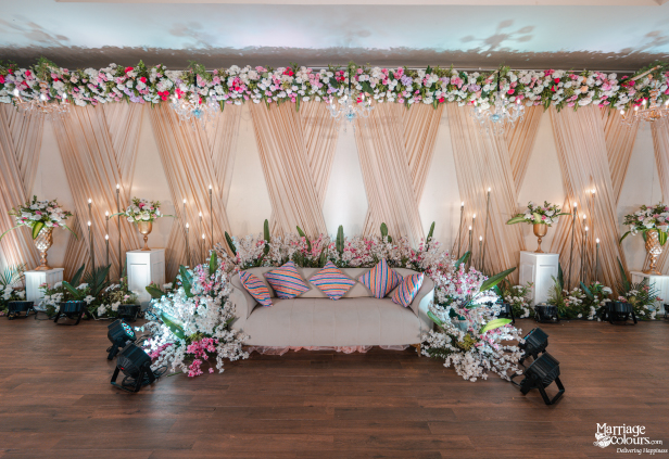 Luxurious wedding stage with a white sofa, bright floral arch, peach drapes, and ambient lighting