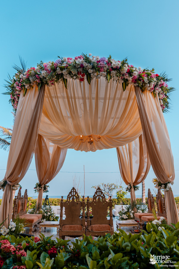 Beautiful outdoor wedding mandap with a floral canopy, peach drapes, and the ocean visible beyond.