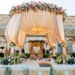 Elegant outdoor wedding mandap with a peach draped canopy, beautiful flowers, and traditional seating.