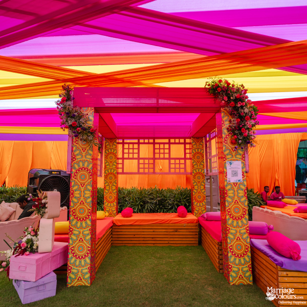 A colorful event seating structure with patterned fabric pillars, orange daybeds, and bright draped ceiling.
