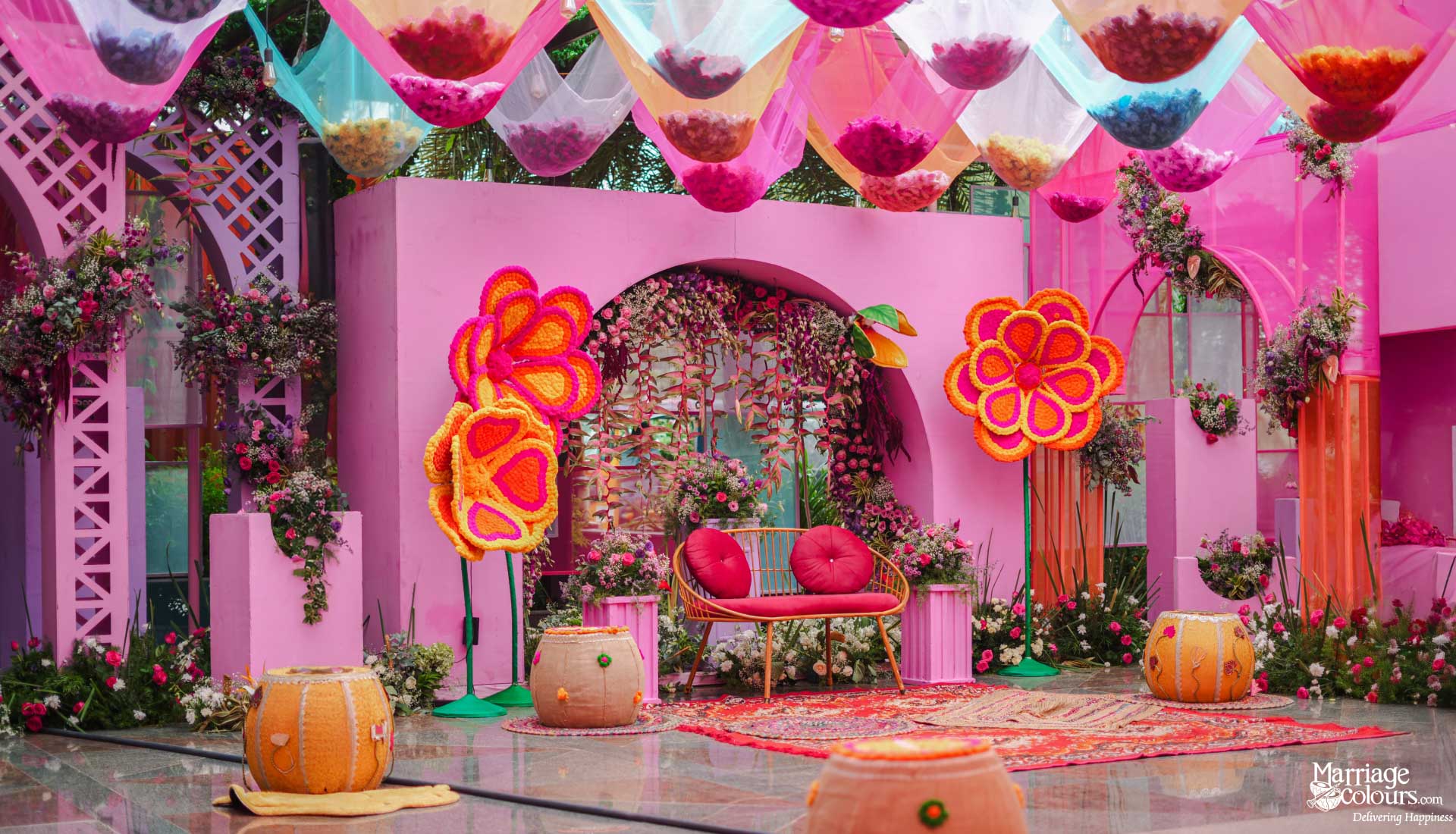 A vibrant pink Mehndi stage with oversized floral decorations and hanging colorful fabrics.