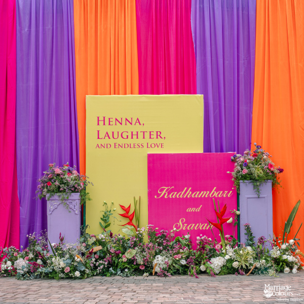 A vibrant event photo backdrop with purple, pink, and orange drapes and floral arrangements.