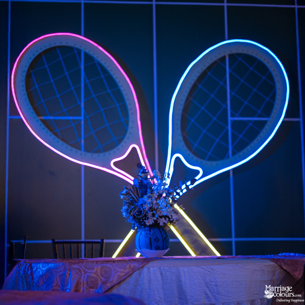 Two large, crossed tennis rackets illuminated by pink and blue neon lights over an event table.