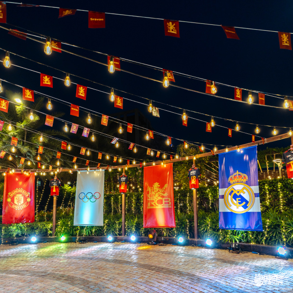 A nighttime outdoor event space decorated with flags of global sports teams and string market lights.