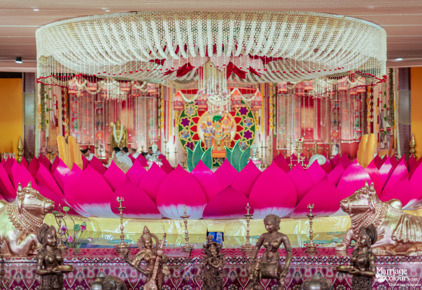 A traditional wedding mandap featuring a giant pink lotus, a floral chandelier, and bronze deity statues in the foreground.