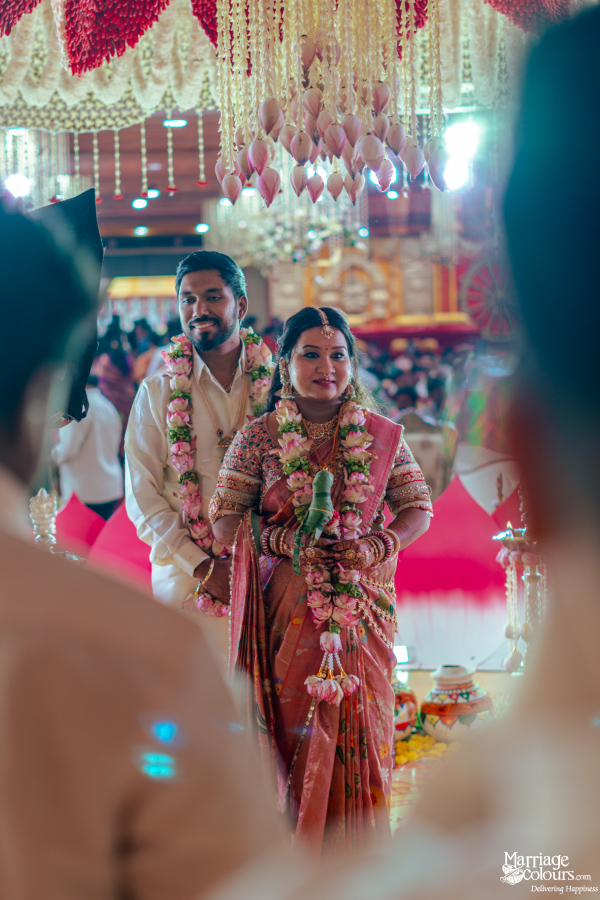 A bride and groom smiling in traditional Indian wedding attire and lush white and pink floral garlands.