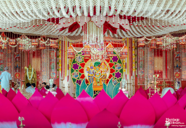 A wide shot of a traditional mandap with a pink lotus foreground, gold statues, and a dense floral chandelier.