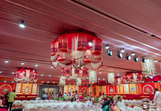 A vast indoor event space with rows of white chairs beneath traditional red and orange elephant-patterned hanging lamps.