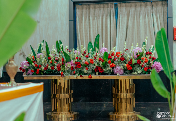 A long, lush floral arrangement of red roses, pink hydrangeas, and white snapdragons on a golden pedestal table.