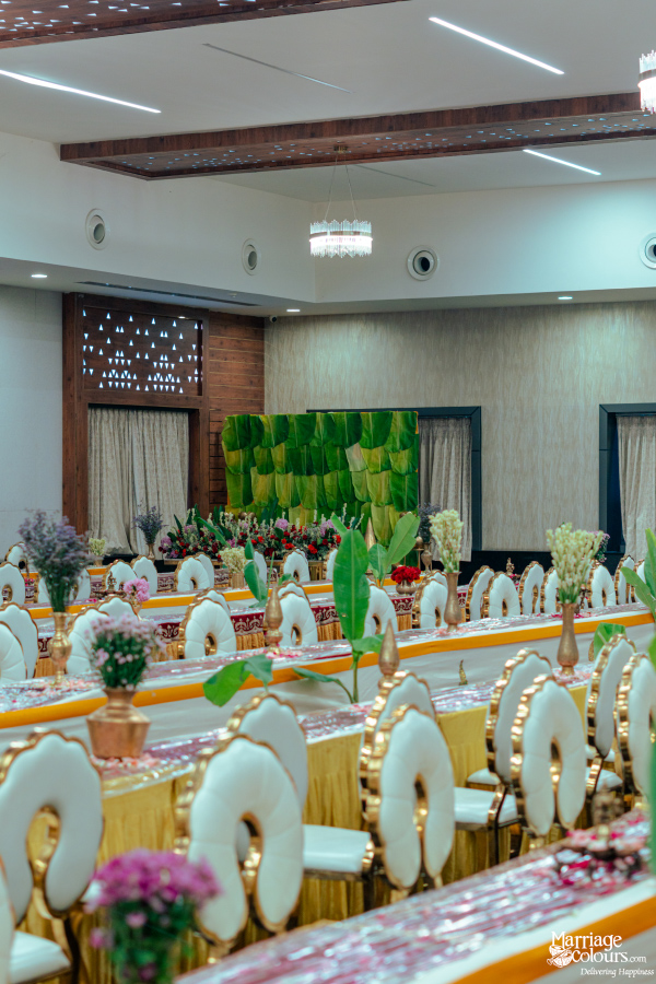 A long, narrow banquet hall set with white and gold patterned chairs and banana leaf table decor.