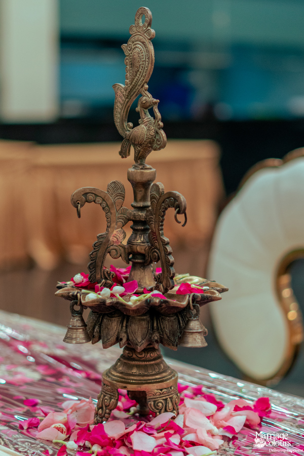 A detailed brass oil lamp with a peacock finial, resting on a table scattered with pink and white rose petals.