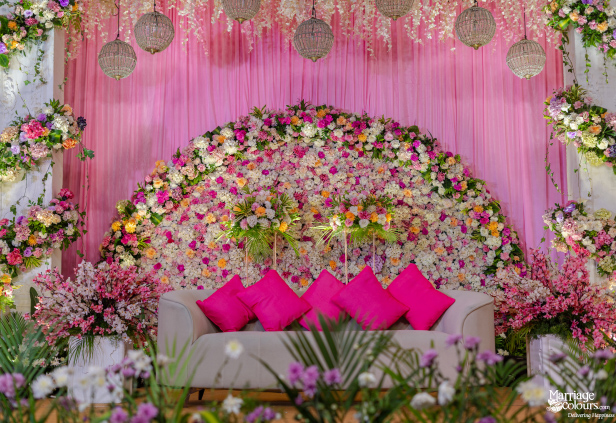 Elegant wedding stage with a white sofa, vibrant floral arch, pink drapes, and hanging crystal globes.