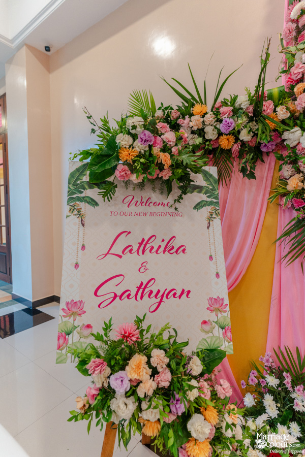 Wedding welcome sign reading "Lathika & Sathyan" surrounded by colorful pink, orange, and purple flowers.
