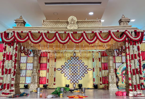 Elaborate golden wedding mandap decorated with dense red and white flower garlands and a central kolam.