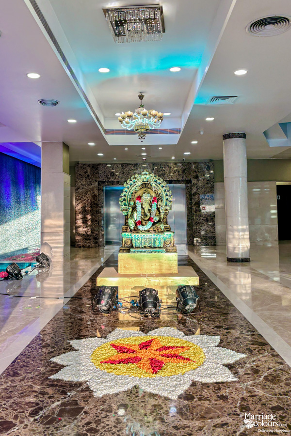 Lord Ganesha statue decorated with flowers, placed on a marble floor with a colorful flower rangoli.