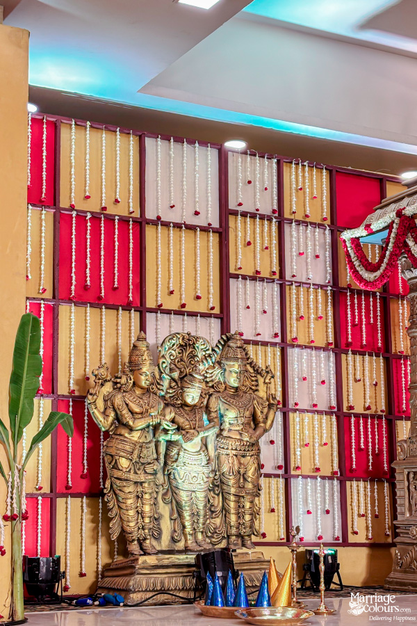 Three golden deity statues in front of a colorful, flower-curtained red, white, and gold wall.
