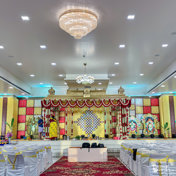 Wedding hall with white chairs, two crystal chandeliers, and a grand red and gold decorated stage.