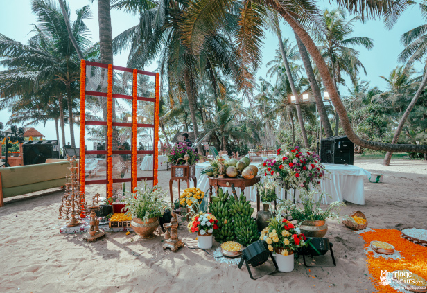 Wide shot of a tropical beach ceremony area with coconuts, bananas, flowers, and traditional lamps.