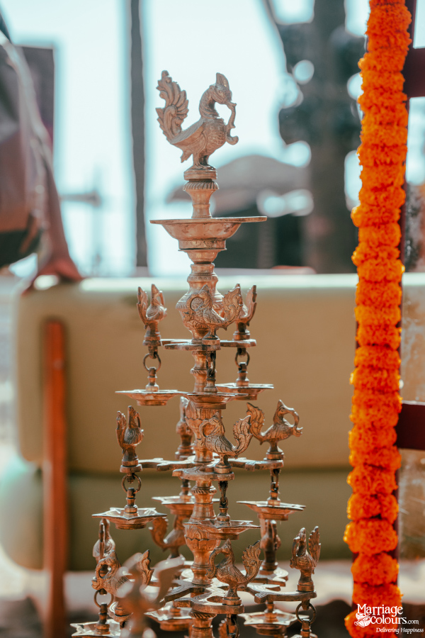 Intricately carved tiered brass oil lamp with a peacock finial, next to a bright marigold garland.