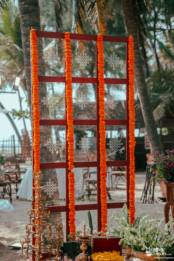 Beach wedding screen with marigold garlands and Kolam stickers on clear panels between palm trees.