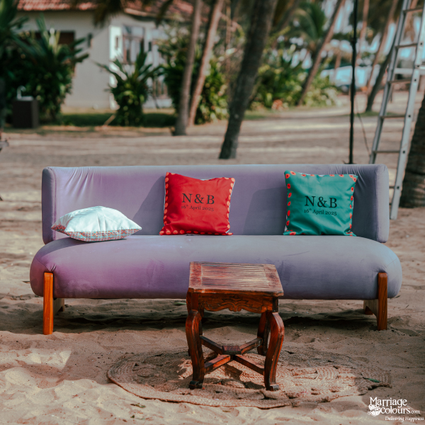 Lavender velvet sofa on the beach with customized red and green cushions under palm trees.